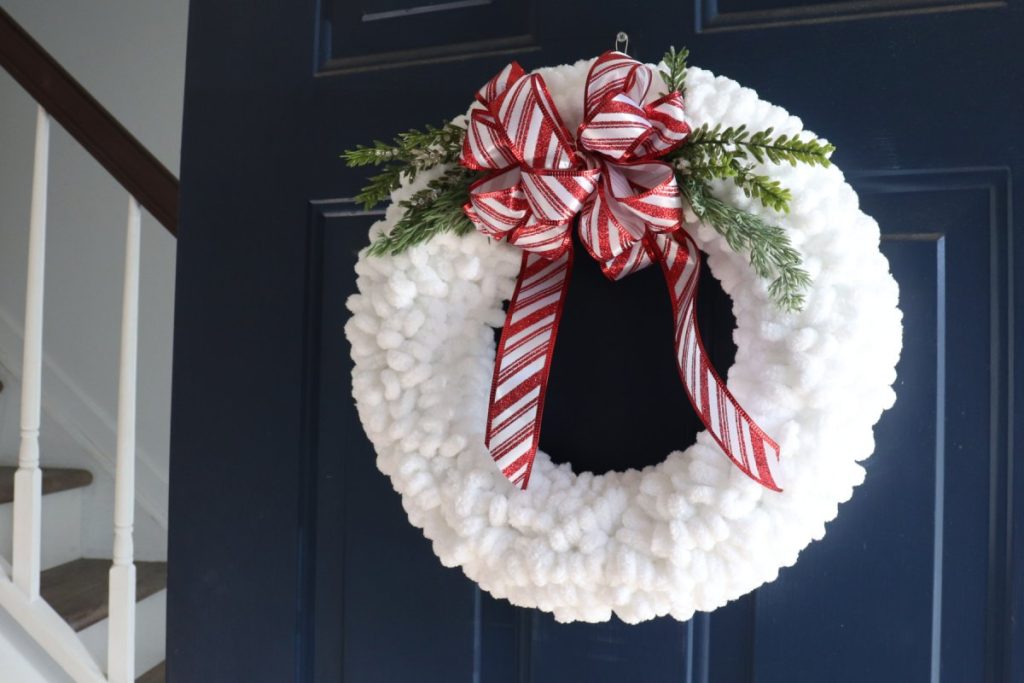 Image contains a wreath made from white loopy yarn, with a red and white striped ribbon bow and faux greenery, hanging on a dark blue door.