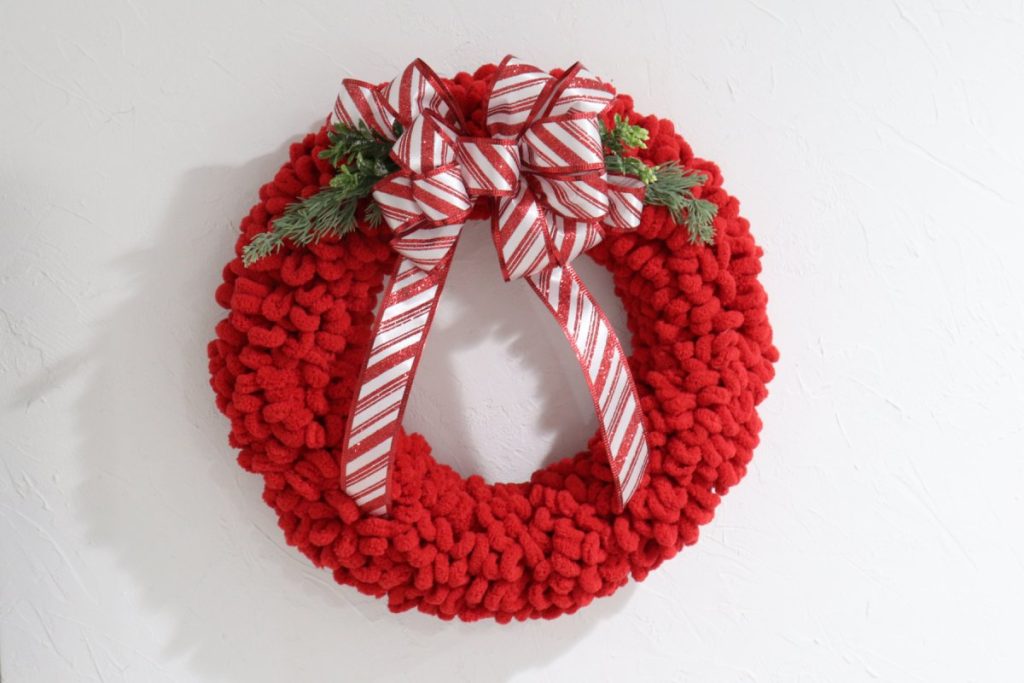 Image contains a wreath made of red loopy yarn, with a red and white striped ribbon bow and faux greenery on a white wall.