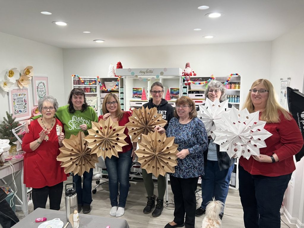 Image contains seven women standing together in Amy’s craft room, smiling while holding their paper bag snowflake and star projects.