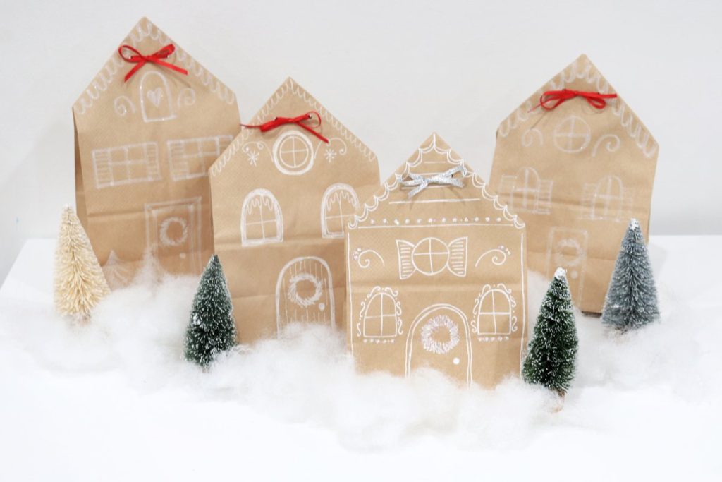 Image contains four brown paper bags decorated with white pen to look like gingerbread houses. They sit among tufts of Poly-Fil with bottle brush trees off to the sides.