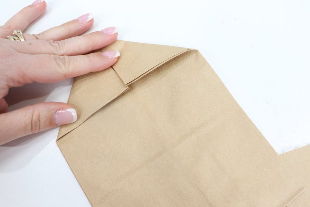 Image contains Amy’s hand folding down the top corners of a brown paper bag to form a triangle.