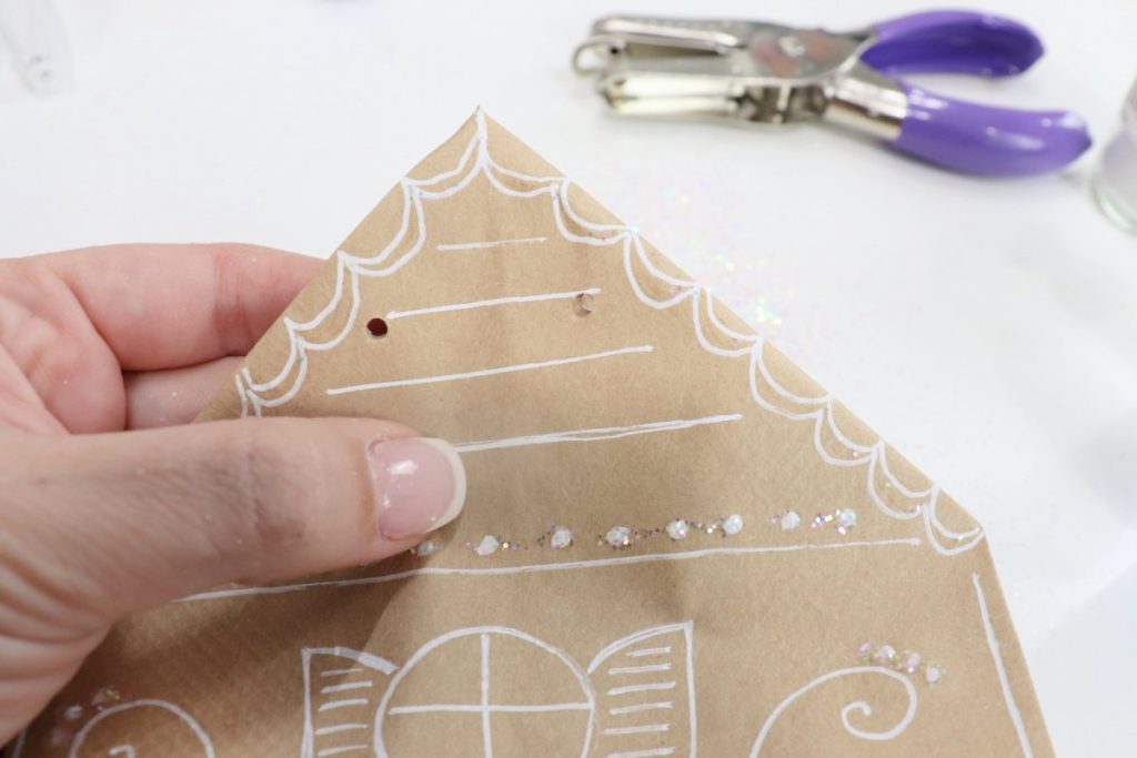 Image contains Amy’s hand holding the top of the paper bag, which has two small holes punched through the layers. A purple-handled hole punch sits off to the side.