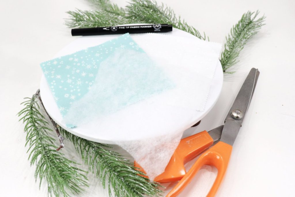 Image contains teal fabric, white batting, a pair of orange handled scissors, and a black fabric marker on a white table with a few pieces of faux pine.