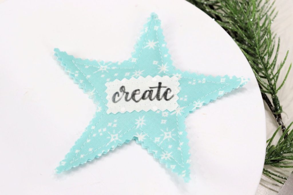 Image contains a teal scrappy star with the word, “create.” It sits on a white table with faux pine branches.