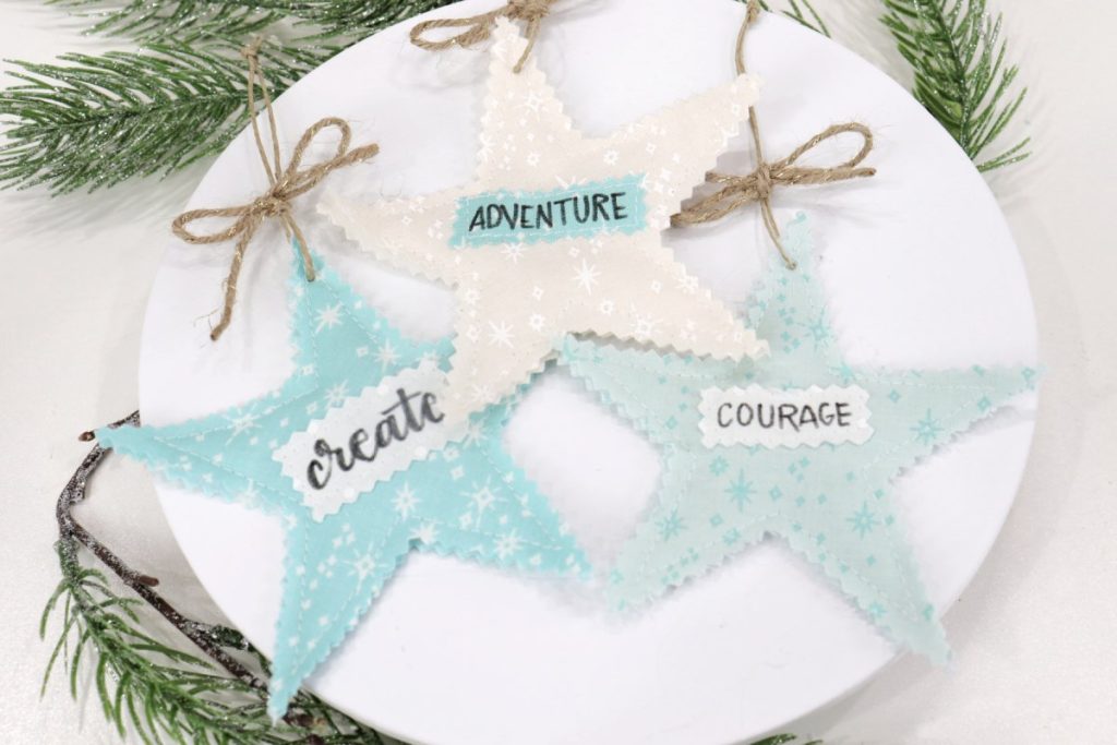 Image contains three scrappy stars; one teal with the word “create,” one white with the word, “adventure,” and one mint with the word, “courage.” They sit on a white tray surrounded by faux pine branches.