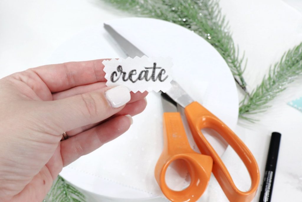 Image contains Amy’s hand holding a small piece of white fabric with the word “create” written in black fabric marker. Below it, a pair of shears and a fabric marker sit on a white table with faux pine branches.