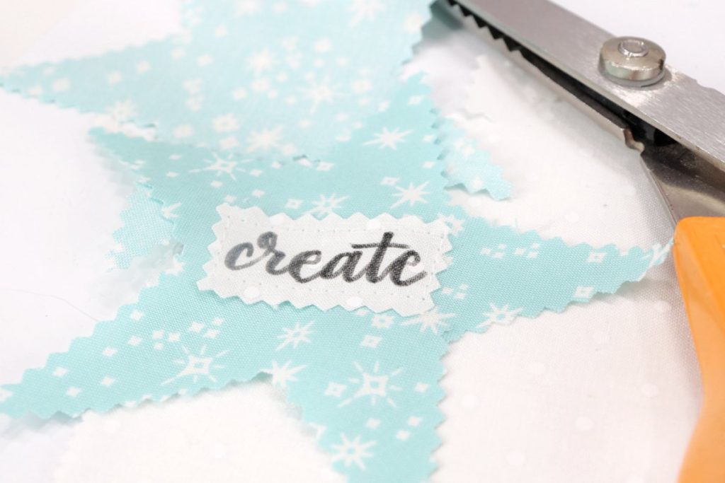 Image contains two teal fabric stars, one with the word, “create” sewn onto it. A pair of scissors sits beside them on a white table.
