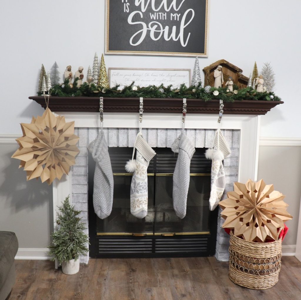 Image contains a whitewashed fireplace with a dark brown mantel. Four stockings hang in front of the fireplace, and there is a large 3D paper star on either side of it. The mantel is decorated with metallic trees and a nativity set.