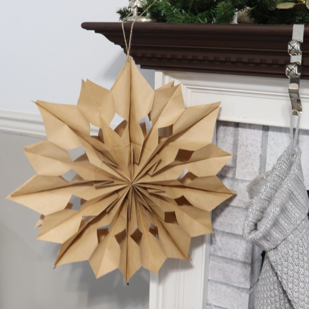 Image contains a 3D paper star made from brown paper lunch bags. It hangs from a fireplace mantel in front of a grey wall.