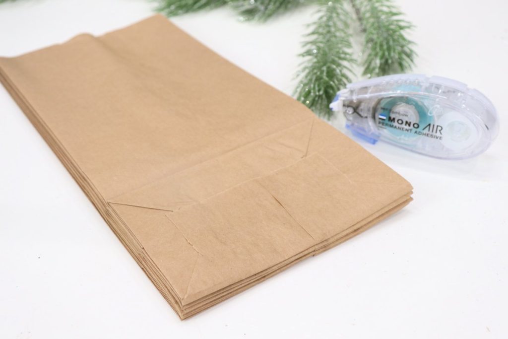 Image contains a stack of 8 brown paper lunch bags glued together. An adhesive runner sits nearby, in front of some faux pine on a white tabletop.