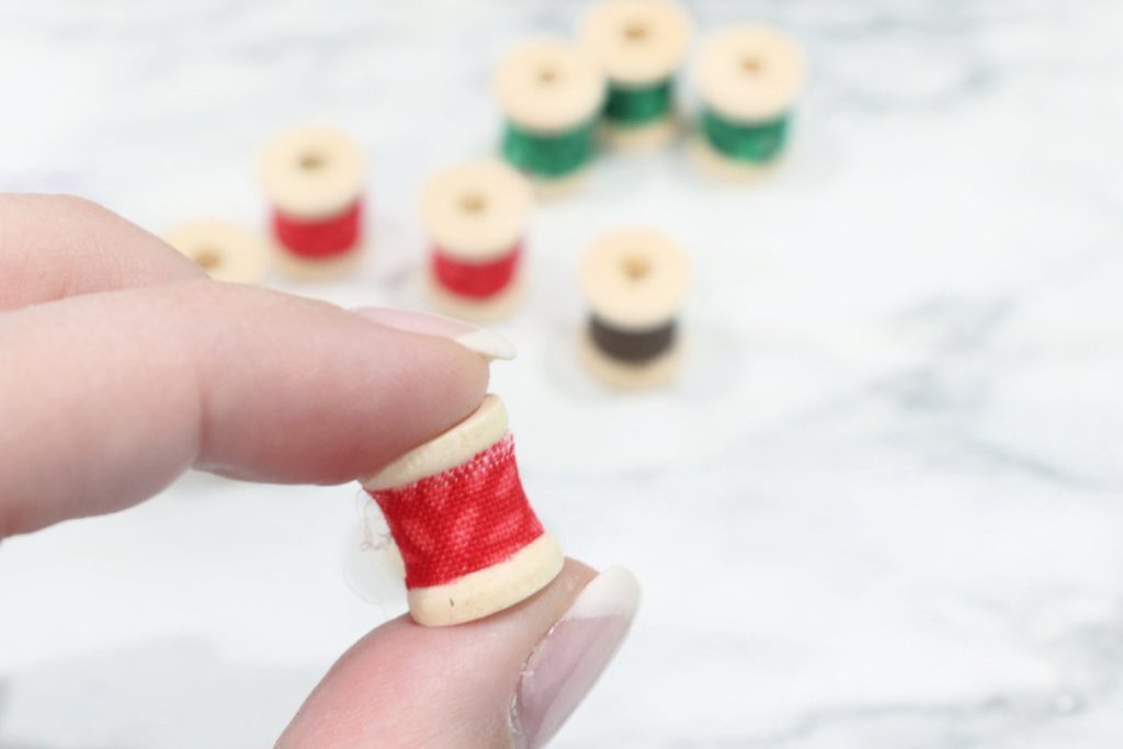 Image contains Amy’s hand holding a 1/2” wooden spool covered with a red strip of fabric. Seven other covered spools sit on a marble countertop in the background, unfocused.