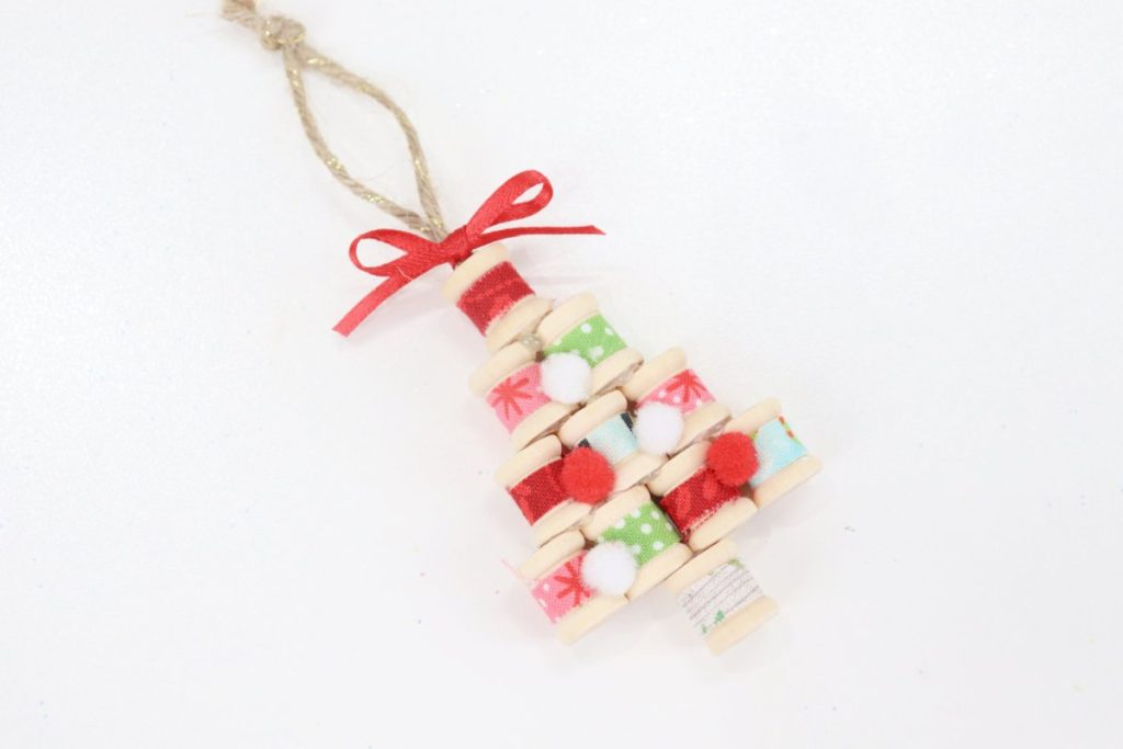 Image contains an ornament made from fabric-covered wooden spools arranged in the shape of a tree. A red bow is tied at the top, and it sits on a white background.