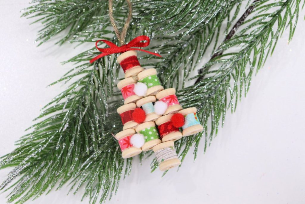 Image contains a tree-shaped ornament made from fabric-covered wooden spools. It has a red ribbon bow at the top, and sits on a faux pine branch.