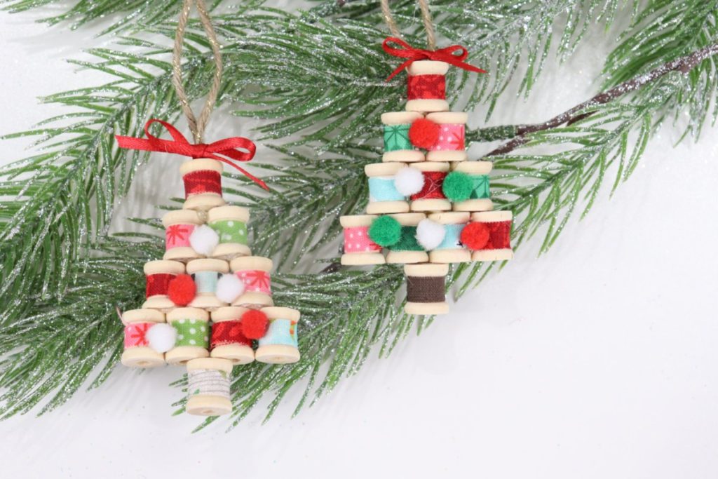 Image contains two ornaments made from fabric-covered wooden spools arranged in tree shapes. They sit on a faux pine branch on a white table.