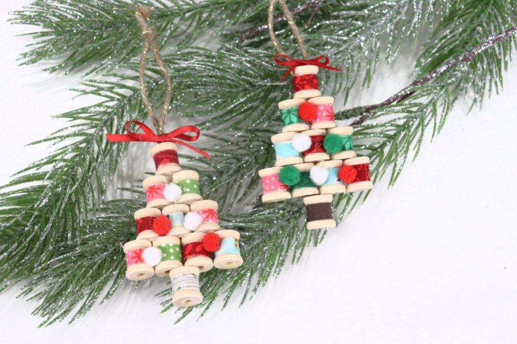 Image shows two ornaments made from fabric-covered wooden spools glued together to form a tree shape. They lay on a faux pine branch that’s sprinkled with glitter.