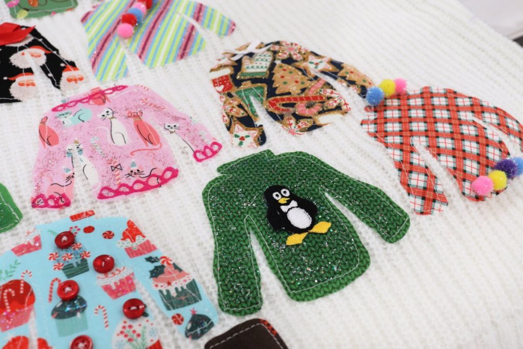 Image contains a closeup of several of the fabric sweaters sewn onto the large sweater. One has been decorated with an iron-on penguin patch, one has five buttons sewn onto it, two are decorated with strings of pom-poms, one has a bow, and one has accents made with fabric paint.