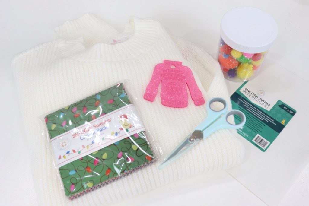 Image contains a white sweater, a pack of 5” fabric squares, a small pink template shaped like a sweater, a blue-handled pair of scissors, a jar of multicolored pom-poms, and fusible interfacing on a white tabletop.