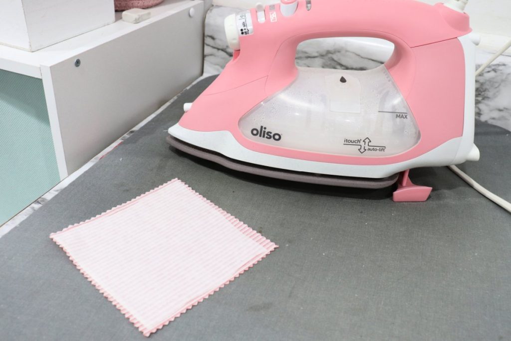 Image contains a 5” square of fabric, wrong side up  on a grey ironing mat. A piece of fusible interfacing lays on top. Behind it is a pink and white Oliso iron.