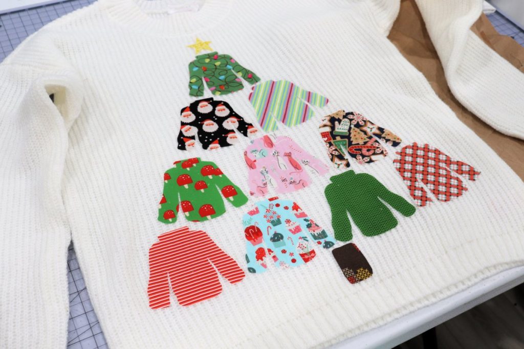Image contains 10 pieces of sweater-shaped fabric arranged in a pyramid with the star shape at the top and the rectangle at the bottom to form a Christmas tree. They are laying on top of a white sweater.