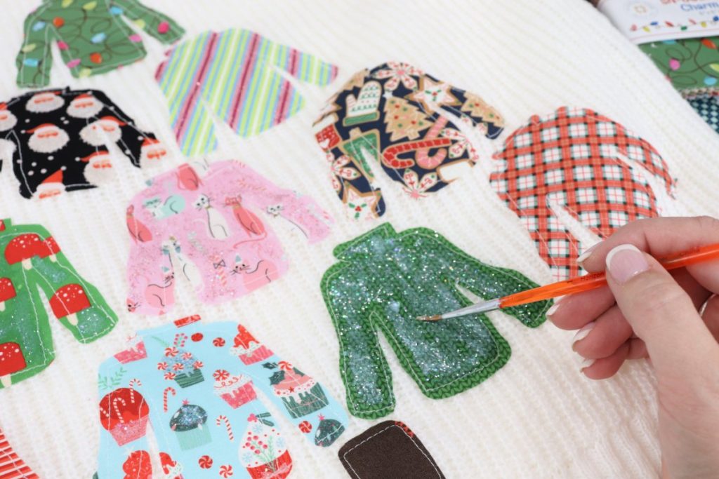 Image contains a white sweater with smaller sweaters cut from a variety of fabrics sewn to the front. Amy’s hand holds an orange paintbrush and applies a layer of fabric glitter to a green sweater.