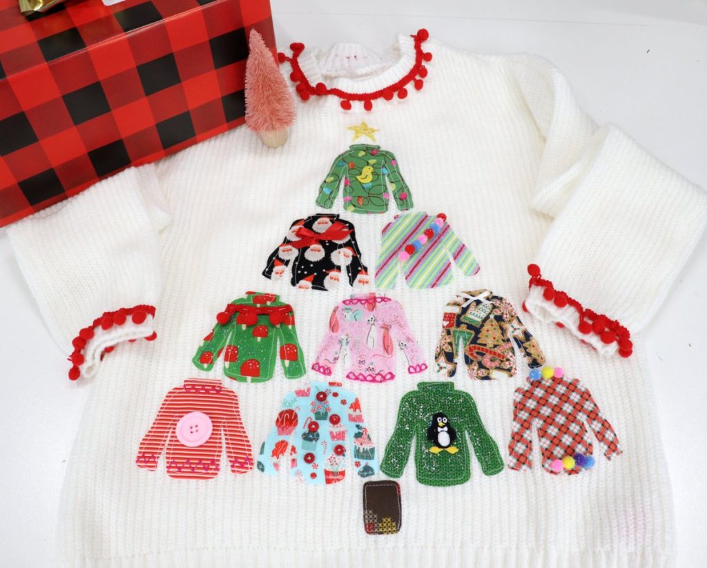 Image contains a white sweater decorated with 10 pieces of fabric cut into sweater shapes and arranged like a Christmas tree. Each sweater is decorated with ribbon, buttons, pom-poms, or fabric paint.