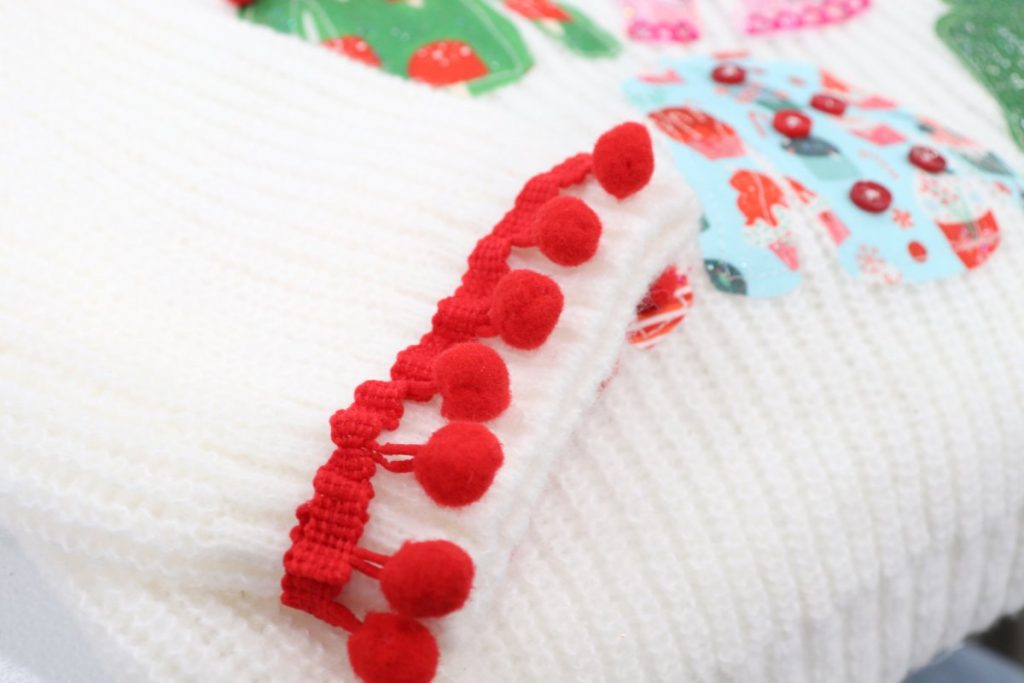 Image contains a closeup view of a white sweater sleeve with a string of red pom poms sewn around the wrist area.