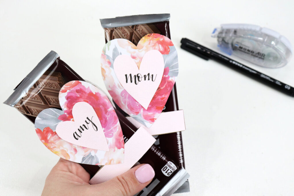 Image contains Amy’s hand holding two chocolate bars with reversible valentines slid onto each one. A marker and an adhesive roll sit in the background on a white table.