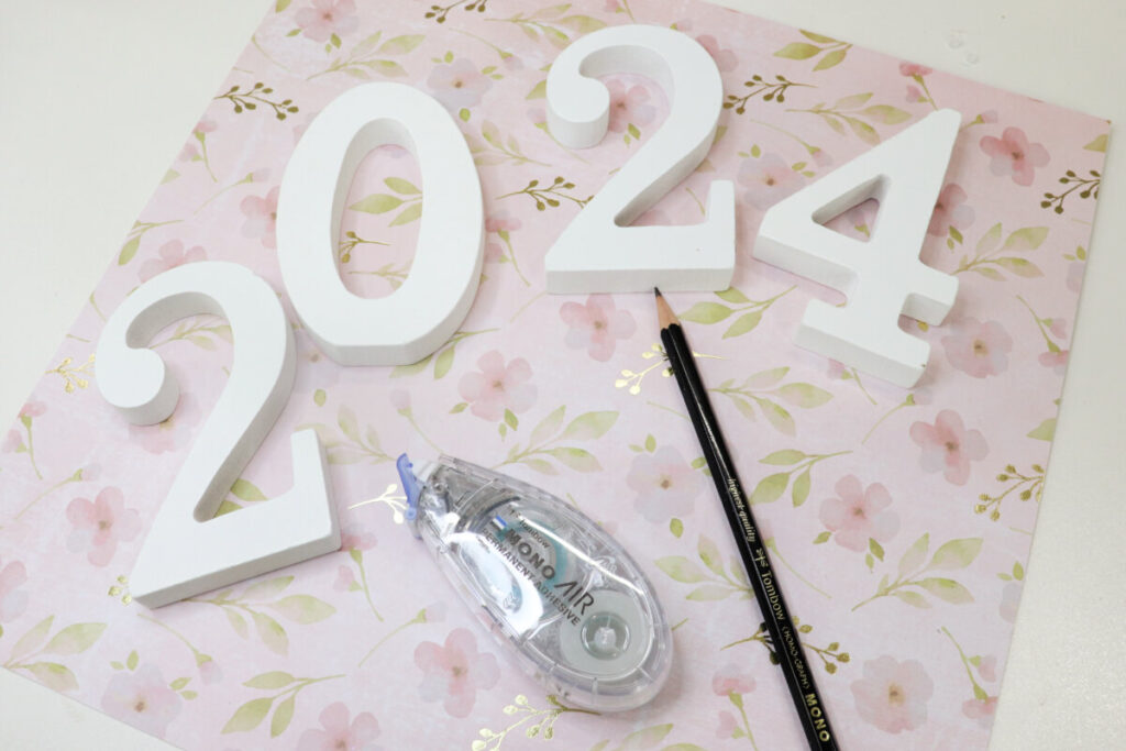 Image contains a piece of pink and gold floral scrapbook paper sitting on a white table. White wooden numbers “2024” sit on top, along with an adhesive tape runner and a sharpened pencil.