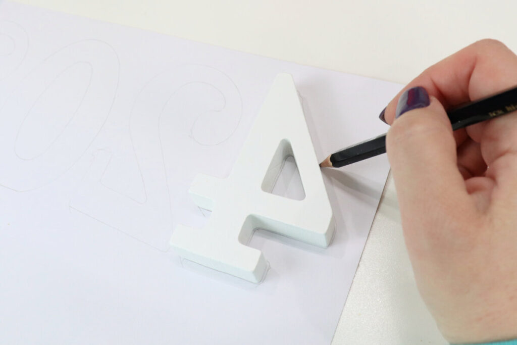 Image contains a white wooden number “4” sitting backward on top of the piece of scrapbook paper. Amy’s hand holds a pencil, tracing around the number.