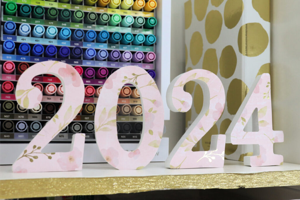 Image contains the decorative “2024” numbers on a white shelf with a colorful marker storage container and a white and gold binder in the background.