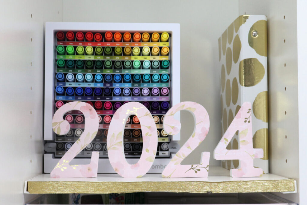 Image contains numbers “2024” covered with pink and gold floral scrapbook paper. The numbers sit on a white shelf in front of a container of multicolored markers and a gold and white polka dot binder.