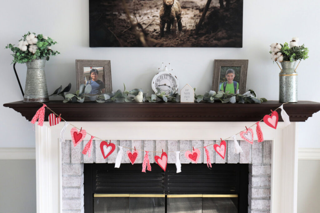 Image contains a white fireplace with whitewashed brick and a dark brown mantel that has been styled for valentine’s day with the banner draped across the front.