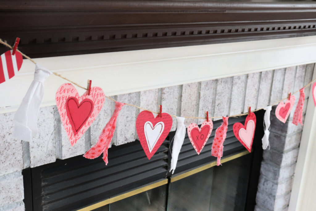Image contains a valentine garland made from cardstock hearts and fabric strips in front of a whitewashed fireplace.