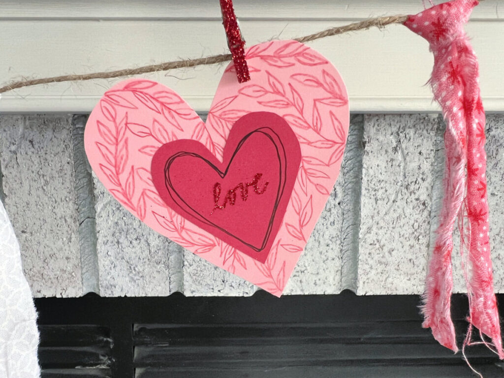 Image contains a close-up view of a pink heart with leafy vines drawn in red colored pencil. A smaller red heart is layered on top with the word “love” written in red glitter script.