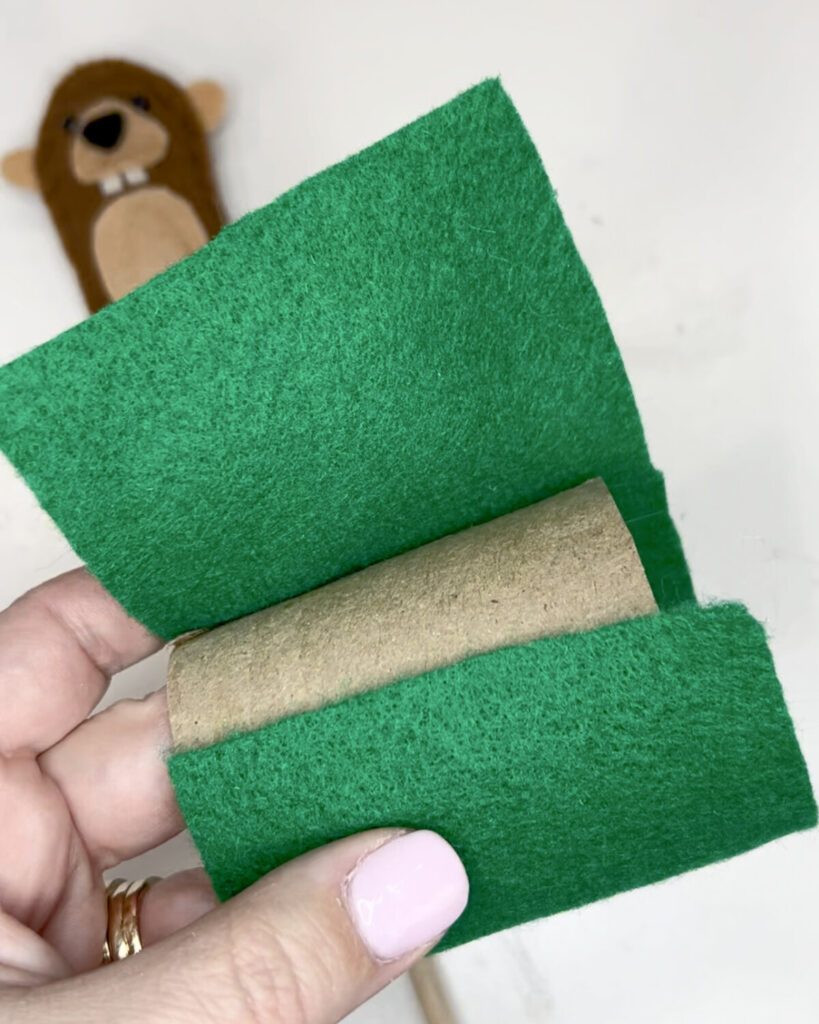 Image contains Amy’s hand holding a paper roll tube and covering it with green felt. The groundhog puppet sits in the background, out of focus.