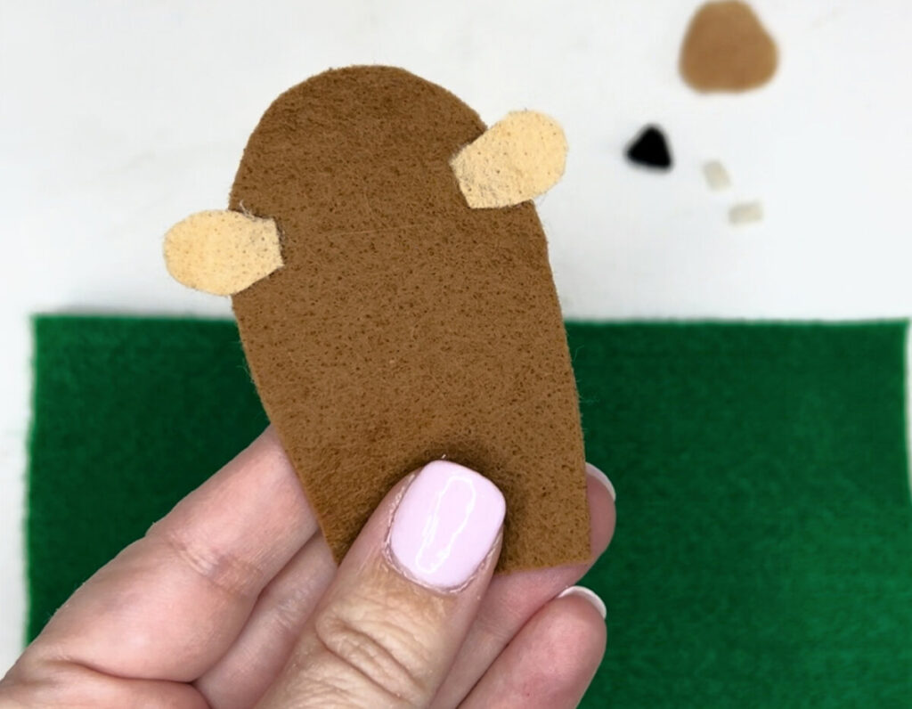 Image contains Amy’s hand holding a piece of brown felt with two small pieces of tan felt glued onto the sides for ears. More felt pieces, including a large green rectangle, sit in the background on a white table.