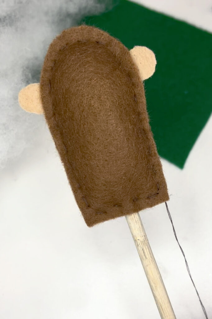 Image contains the plush groundhog body with ears, and a dowel extending from below the body. A piece of green felt and a pile of Poly-Fil sit on a white table behind it.