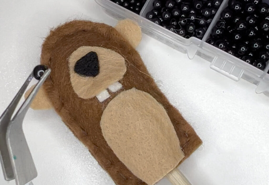 Image contains the plush felt groundhog laying on a white table top next to a box of black beads. A pair of tweezers holds a black bead, ready to place it on the groundhog’s face as an eye.