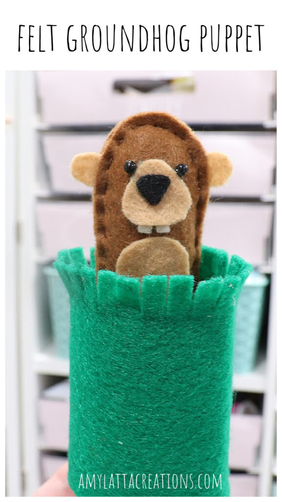 Image contains a plush felt groundhog popping out of a toilet paper tube covered in green felt “grass.” 