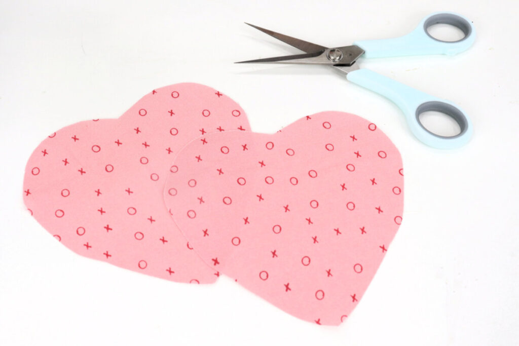 Image contains two hearts cut from pink fabric and a pair of scissors with mint colored handles on a white table.