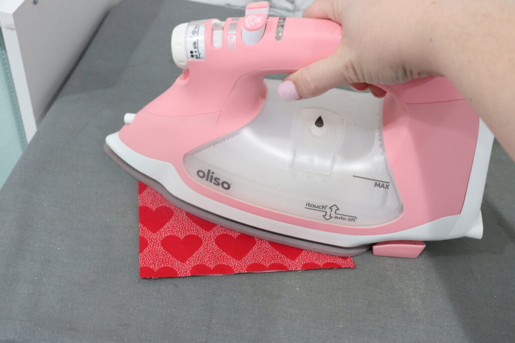 Image contains Amy’s hand holding a pink Oliso iron to press a rectangle of red fabric on a grey mat.