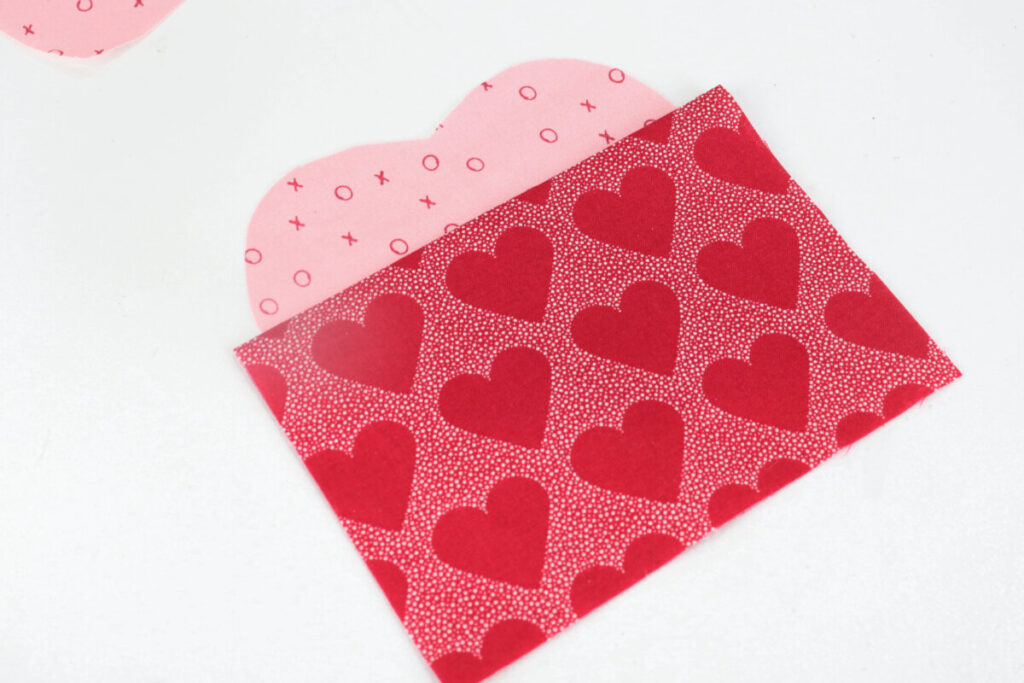 Image contains a pink fabric heart with a folded rectangle of red heart-printed fabric laying on top, covering the bottom portion.