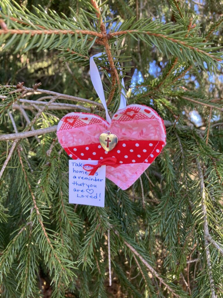 Image contains a red and pink fabric heart hanging in a pine tree.