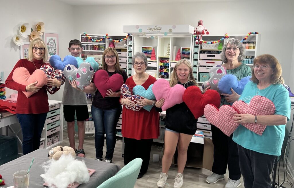 Image contains a group of seven people smiling and holding plush heart-shaped pillows in Amy’s craft room.