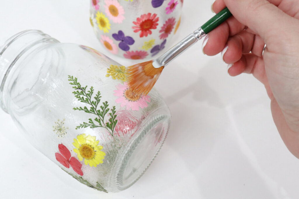 Image contains Amy’s hand holding a paintbrush and applying decoupage formula to a clear jar covered in pressed flowers. A second floral jar sits nearby on a white table top.