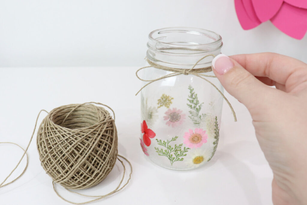 Image contains Amy’s hand tying a twine bow around the top of a clear jar covered in pressed flowers. A roll of twine sits nearby on a white table.