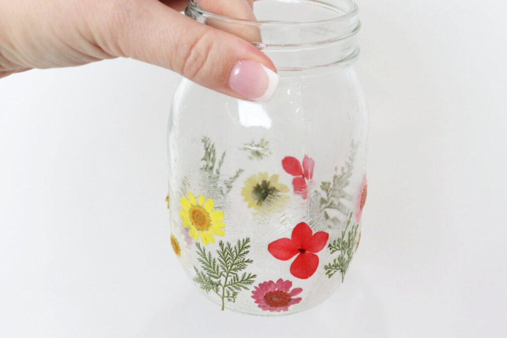 Image contains Amy’s hand holding a jar covered with assorted pressed flowers and leaves against a white background.