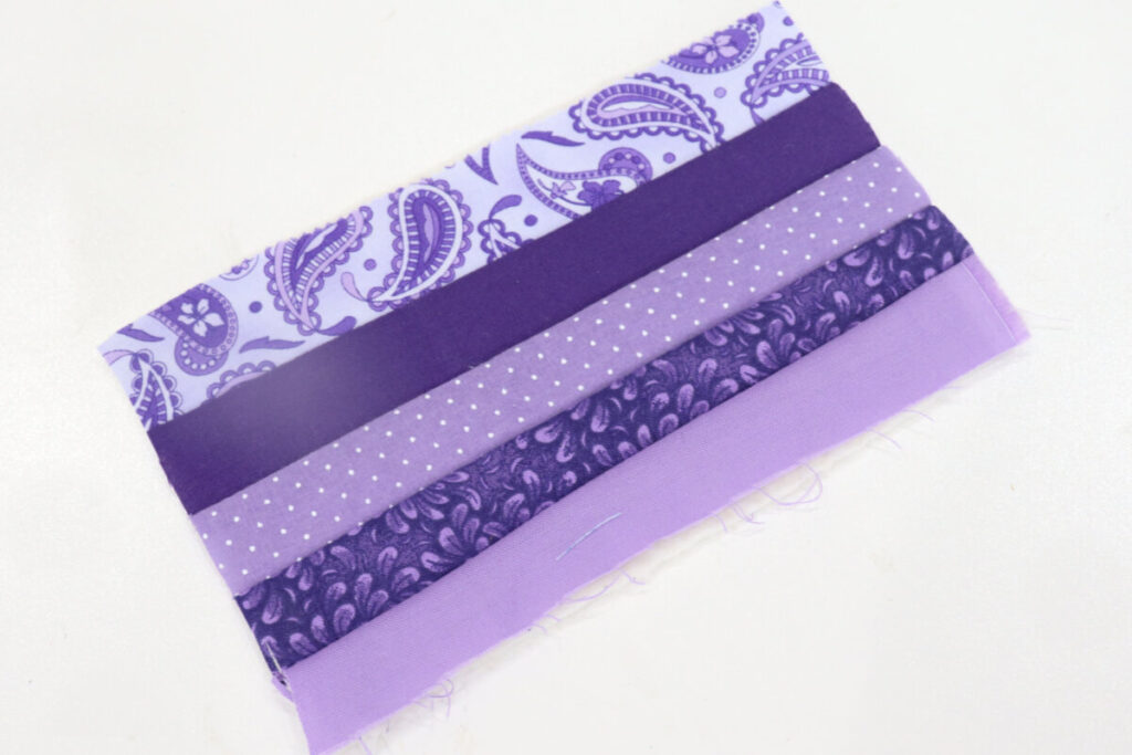 Image contains five purple fabric strips of various patterns sewn together to form a rectangle.