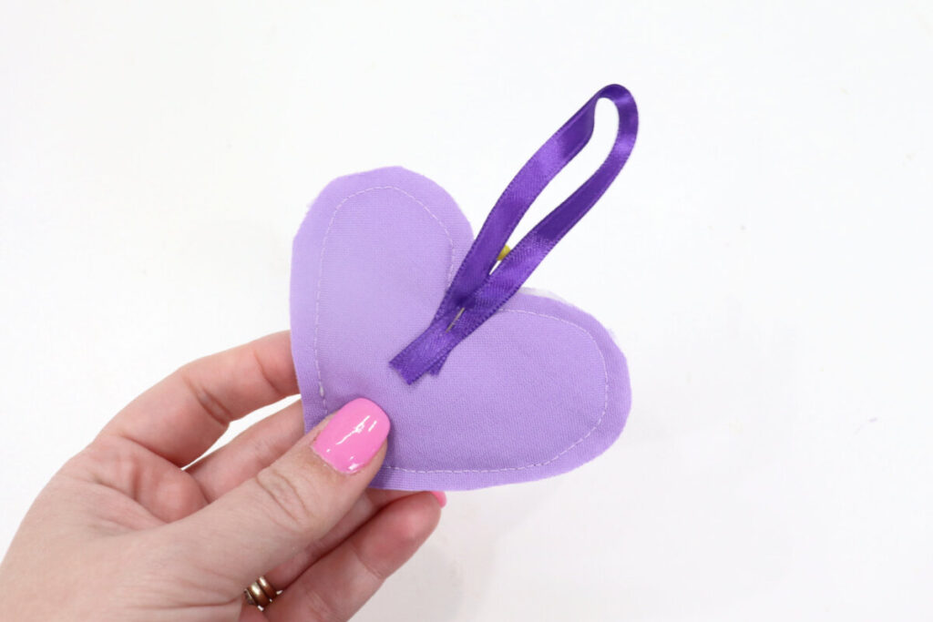 Image contains Amy’s hand holding the fabric heart with the back facing up and a purple ribbon loop pinned in place.