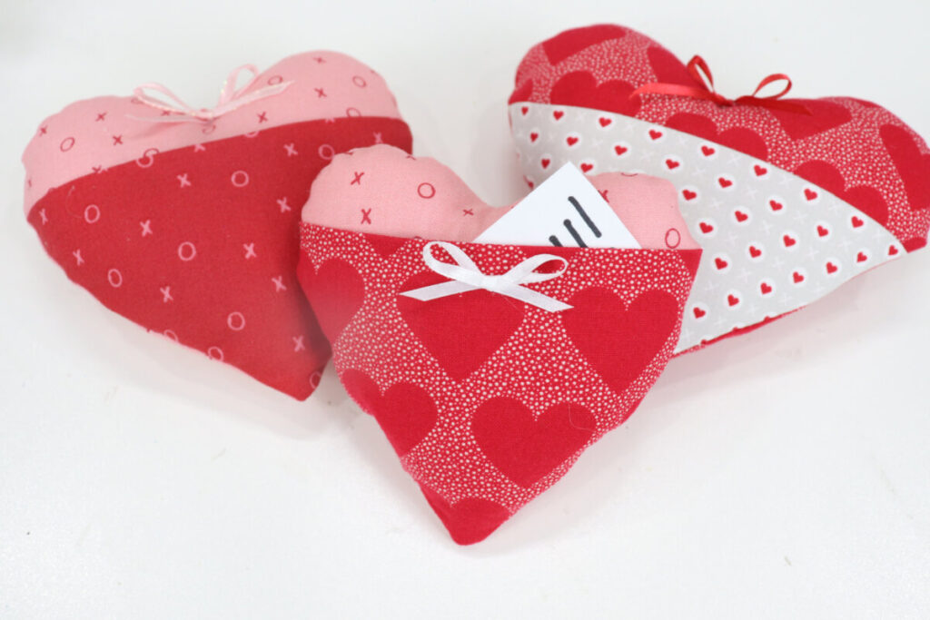 Image contains three plush hearts made of assorted red, pink, and neutral fabrics with a pocket on the front.